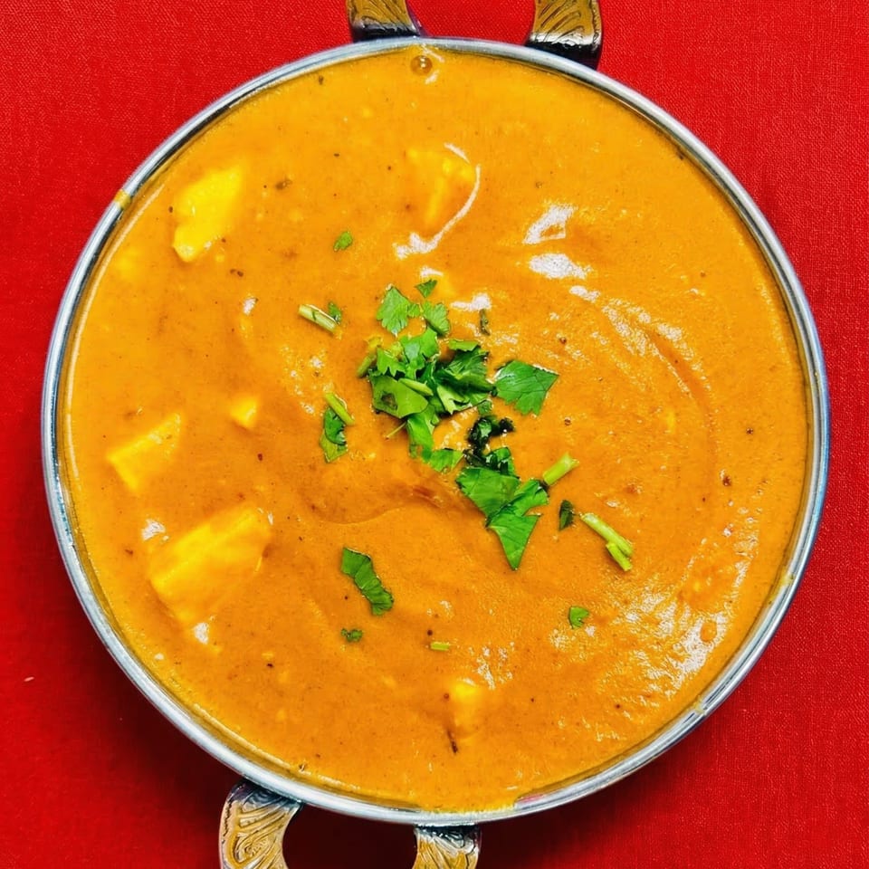 Indian paneer butter masala, a creamy orange curry with cheese cubes and cilantro, in a metal bowl.