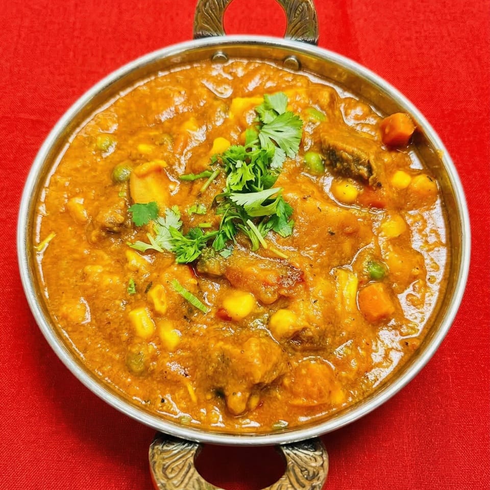 Indian curry with meat, vegetables, and fresh cilantro in a metal bowl on a red tablecloth.