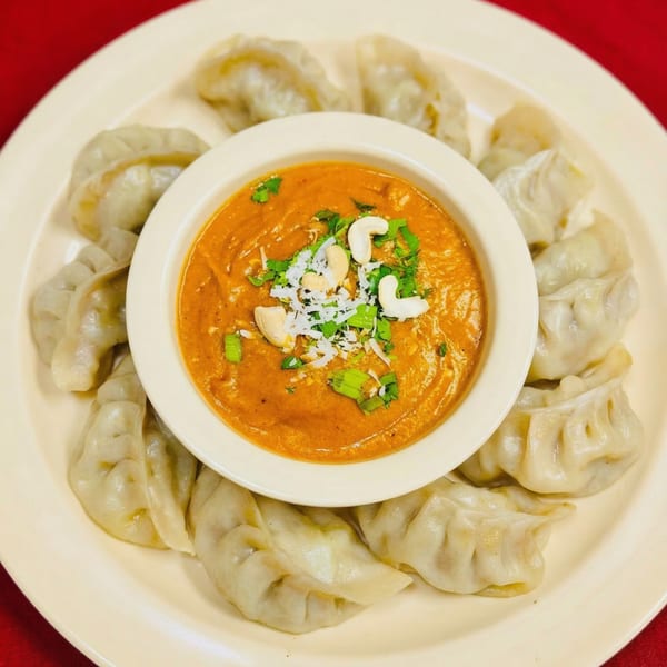 A plate of Indian momos, steamed dumplings, surrounding a bowl of orange dipping sauce garnished with cashews and herbs.